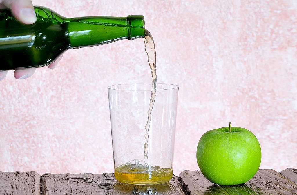 Imagen de una botella vertiendo sidra en un vaso con una manzana al lado